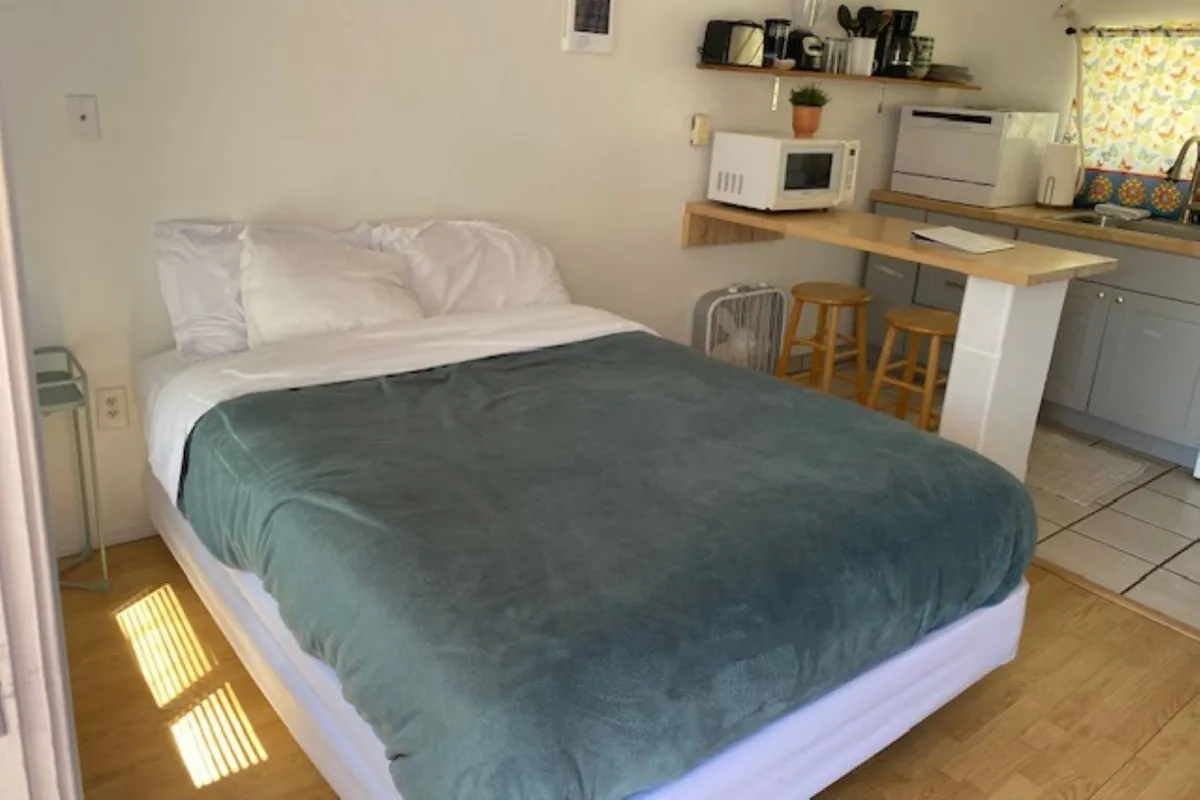 This image showcases the core of the studio: a comfortable bed positioned near a compact kitchenette, with bar stools offering a spot for casual dining or conversation. The space is bright and inviting, suggesting a seamless blend of living, dining, and kitchen areas. The updated furnishings and décor reinforce the unit’s fresh coastal vibe.