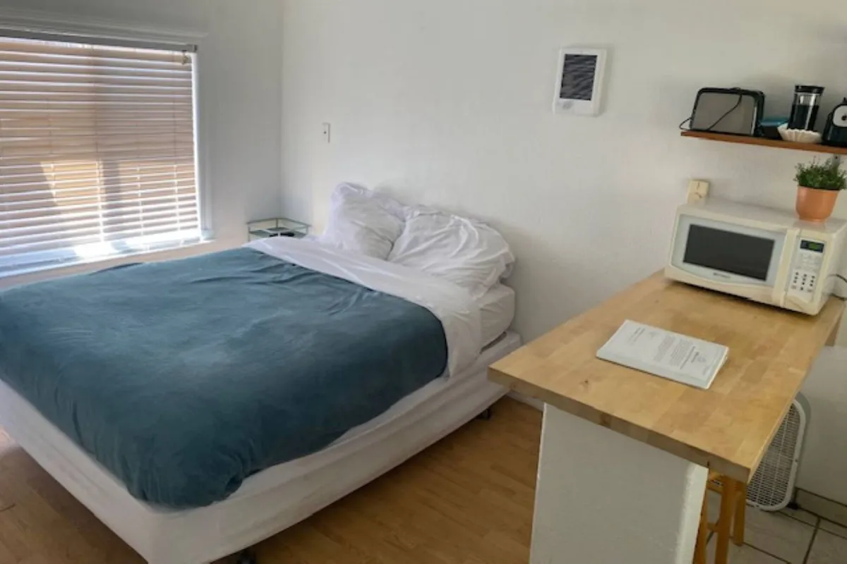A different angle on the studio, this shot features the bed, compact kitchenette, and a sunlit window with blinds. The relaxed scene suggests quiet mornings, reading in bed, or enjoying a cup of coffee in natural light.