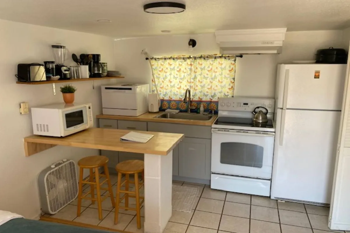Focusing on the kitchen area, this image features a well-equipped countertop, appliances, and stools beneath a window adorned with a colorful curtain. The scene feels warm and cozy, ideal for preparing meals or enjoying a morning coffee.